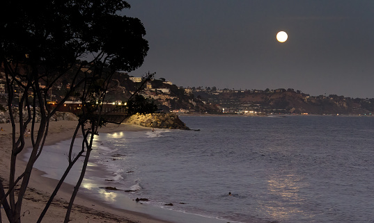Night landscape of Pacific Palisades on the California coast, with full moon rising and reflections on the ocean water.