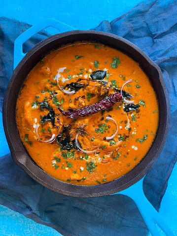 Stock photo showing elevated surface view of turquoise blue cooking pan filled with Indian butter chicken, chicken tikka curry sauce. Butter chicken breast chunks sprinkled with red onion and mustard seeds.