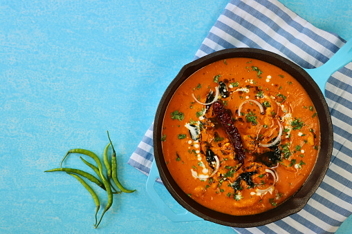 Stock photo showing elevated surface view of Indian butter chicken, like chicken tikka masala in turquoise blue cooking pan. Butter chicken breast chunks sprinkled with red onion and mustard seeds, served with lachha paratha (layered flatbread)