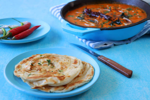 Stock photo showing elevated surface view of Indian butter chicken, like chicken tikka masala in turquoise blue cooking pan. Butter chicken breast chunks sprinkled with red onion and mustard seeds, served with lachha paratha (layered flatbread), red chillis. imagen de pollo de mantequilla india / curry tikka servido en sartén azul turquesa lleno de grandes trozos de carne de pechuga de pollo en salsa de curry con guarnición de rodajas de cebolla roja y semillas de mostaza, lachha paratha (pan plano en capas)