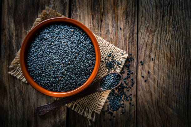 Overhead view of a clay bowl filled with black sesame seeds shot on rustic wooden table. A wooden spoon is beside the bowl and some seeds are scattered on the table. Predominant colors are black and brown. High resolution 42Mp studio digital capture taken with Sony A7rII and Sony FE 90mm f2.8 macro G OSS lens