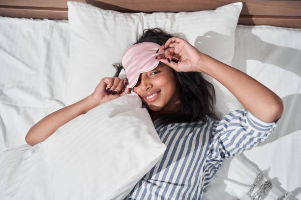 Woman laying in her bed in striped pajamas and sleep mask Cute brunette multiracial woman laying in her bed in striped pajamas and sleep mask, top view, Girl looking flirty at the camera and relaxing. Stock photo sleep eye mask stock pictures, royalty-free photos & images