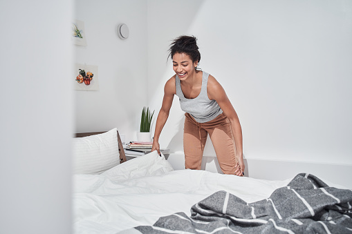 Young beautiful concentrated woman making her bed early in the morning while looking happy. Woman adjusting blanket at the bed with happy smile. Stock photo