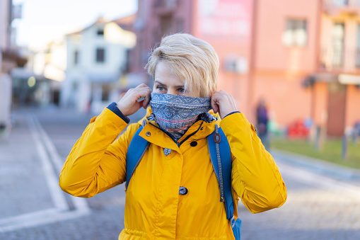 Portrait of beautiful blonde woman, wearing a neck gaiter and yellow jacket on the street.