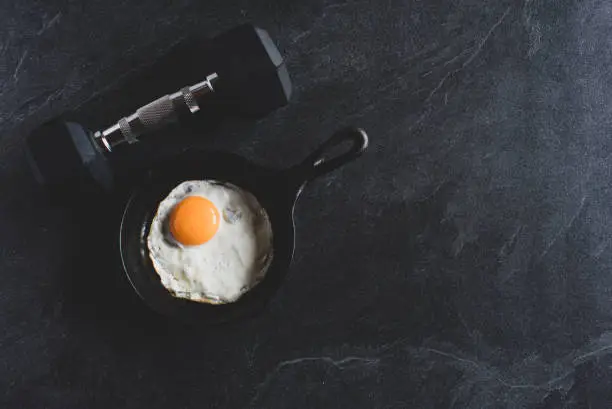 background image for sports concepts with a dumbbell and a fried egg sunny side up in iron cast pan on dark background with an overhead view