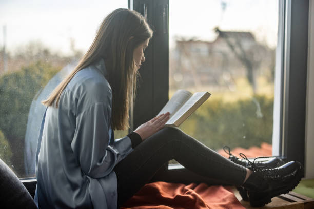 istock Young woman sitting on bench near window and reading book 1298618429