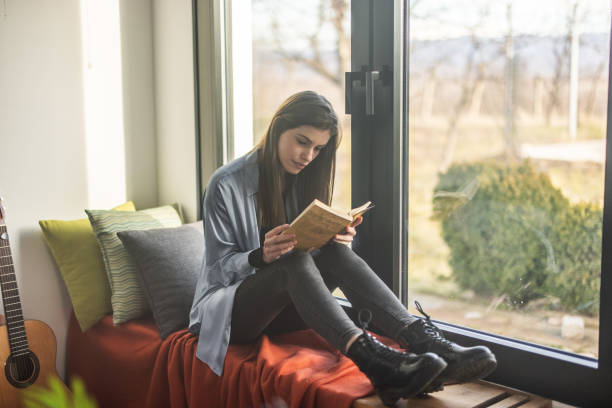 istock Young woman sitting on bench near window and reading book 1298618436