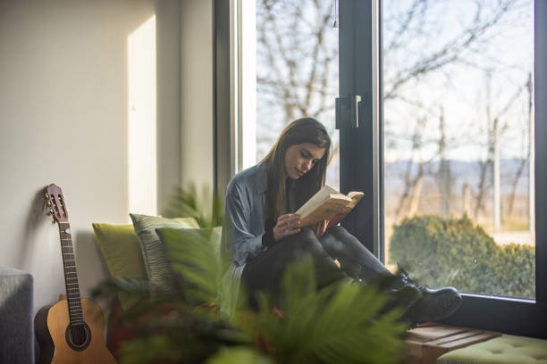 istock Young woman sitting on bench near window and reading book 1298618447