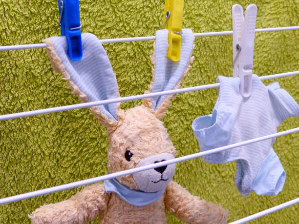 The photo shows a cute plush bunny after washing. To dry, the hare and its clothes were fastened with clothespins on a hills hoist. In the background hangs a terry towel.