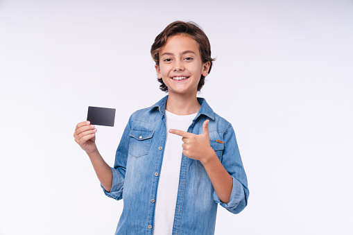 Smiling boy pointing at credit card in casual attire isolated over white background