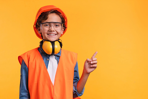 Small caucasian boy construction worker in uniform pointing at copy space isolated over yellow background
