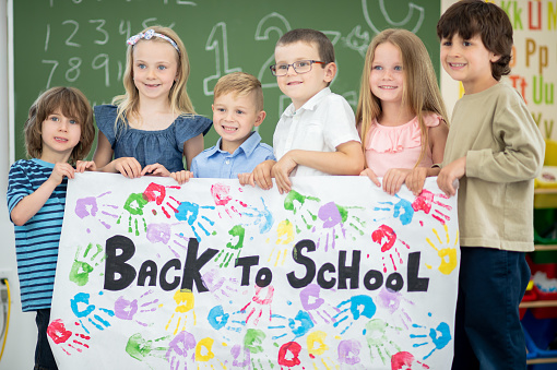 A portrait of a group of elementary age children standing in front of a Blackboard in class. They are holding a banner that has \