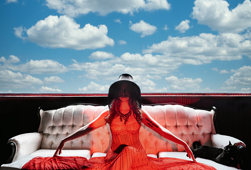 Conceptual composite image of Female artist performer relaxing on the sofa. She is wearing a lamp shade on her head as a part of her performance. Black cat in the corner is looking at her. Cloudy sky in the background.