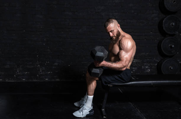 istock Young active strong sweaty fit muscular man with big muscles sitting on the bench and doing biceps workout with dumbbells in the gym as hardcore cross training 1298645885