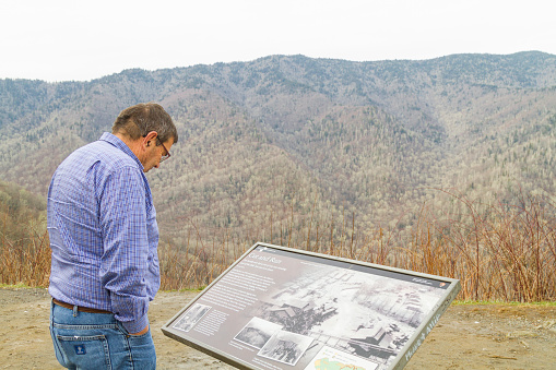December 15, 2020 - Pigeon Forge, Tennessee, USA: A tourist male reading a sign board with information about the nature and animals around Great Smoky Mountain Valley, on back, an amazing landscape view with a winter forest in a cold evening.