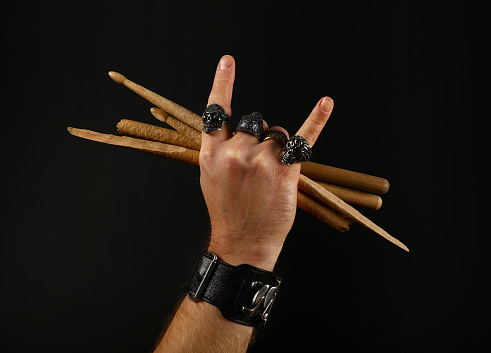 Close up man hand with devil horns rock gesture sign, metal rings and bracelet holding two crossed broken drumsticks over black background, side view