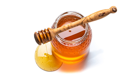 Overhead view of an open honey jar wit a wooden honey dipper on it isolated on white background. Predominant colors are gold and white. The composition is at the right of an horizontal frame leaving useful copy space for text and/or logo at the left. High resolution 42Mp studio digital capture taken with Sony A7rII and Sony FE 90mm f2.8 macro G OSS lens