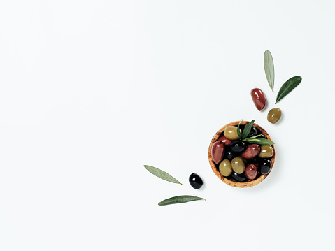 Olives tree leaves and fruits on white background. Small bowl with green, black and red kalamata olives, top view or flat lay. Olives isolated on white with copy space