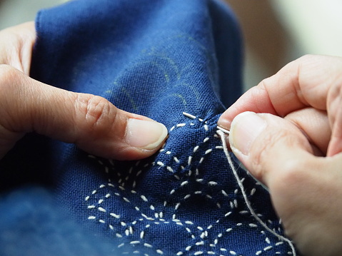 Close up needle in woman's hands sewing running stitch in blue fabric. Traditional Japanese sewing pattern call \