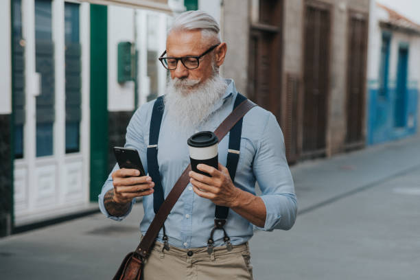 istock an older man in hipster clothes and glasses and a long white beard drinks coffee while walking down the street and looks at his mobile phone focus on head 1298709525
