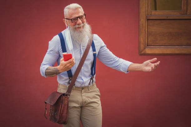 an older man in hipster clothes and glasses and a long white beard walks around talking on the phone focus on head stock photo