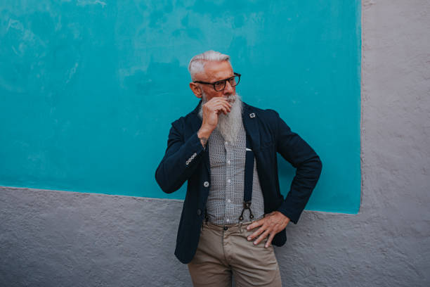 an older man in hipster clothes and glasses and a long white beard poses on a blue wall stock photo