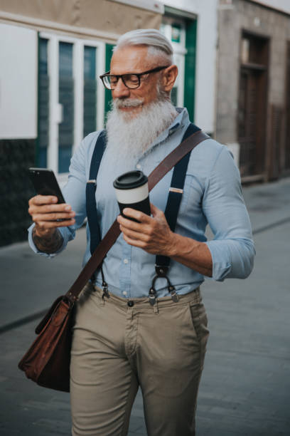 istock an older man in hipster clothes and glasses and a long white beard drinks coffee while walking down the street and looks at his mobile phone focus on head 1298709595