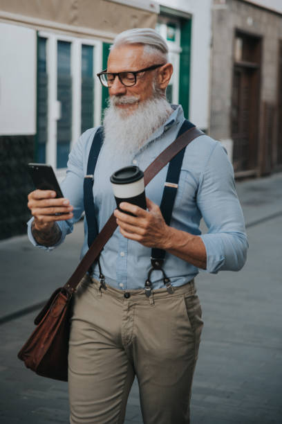 an older man in hipster clothes and glasses and a long white beard drinks coffee while walking down the street and looks at his mobile phone focus on head stock photo