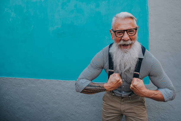 an older man in hipster clothes and glasses and a long white beard poses on a blue wall stock photo