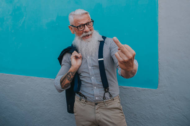 an older man in hipster clothes and glasses and a long white beard poses on a blue wall focus on head stock photo