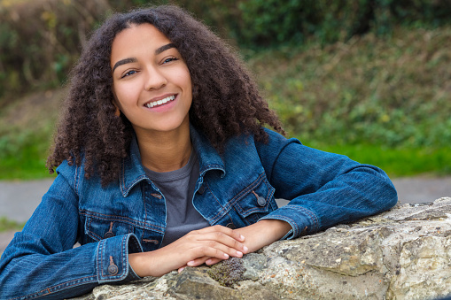 Outdoor portrait of beautiful happy mixed race African American girl teenager female child smiling with perfect teeth wearing denim jacket leaning on a stone wall