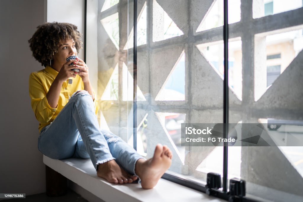 Young woman drinking coffee relaxing on window sill at home Hope - Concept Stock Photo