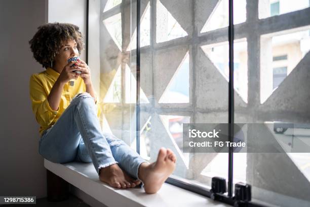 Young Woman Drinking Coffee Relaxing On Window Sill At Home Stock Photo - Download Image Now