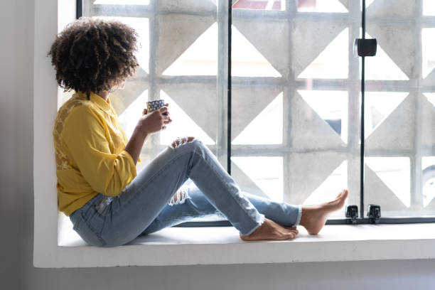 istock Young woman drinking coffee relaxing on window sill at home 1298715519