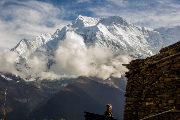 Spectacular view of Mount Annapurna II (7.937 m)and a Nepali man staring smiling in the foreground. A typical nepali stone building is close to him. \nThe picture was taken in Upper Pissang, Nepal, during the Annapurna Round Trek in september 2018.