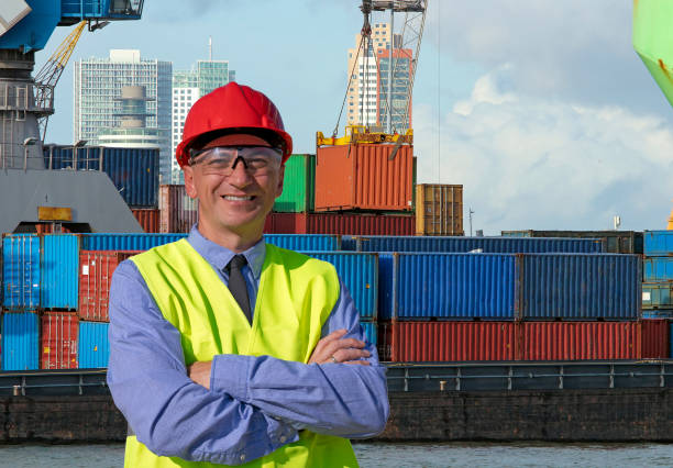 istock Smiling Worker in Red Hardhat and Yellow West Standing Near Container Terminal 1298733539