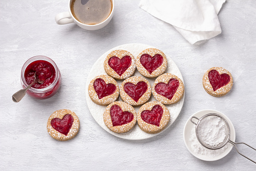 Homemade shortbread spicy cookie with red hearts with jam on white plate and a cup of coffee  on gray textured background, for valentine's day. flat lay
