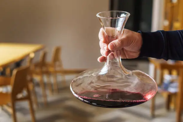 Photo of Close up on hand of unknown caucasian man holding decanter with red wine in the hall at home or restaurant with copy space