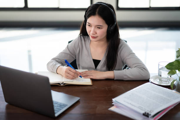 istock Young Asian girl university student wears wireless headphones write on the notebook to study language online watch and listen to the lecturer, webinar via video call e-learning at home, distance education 1298770805