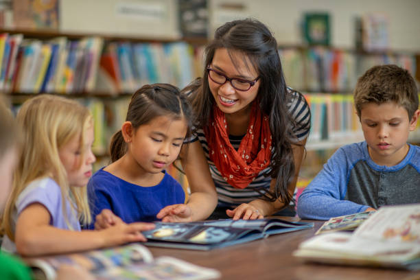 This book here is very interesting A female teacher of Asian ethnicity is helping her multi ethnic group of students with a book to read. They are all dressed casually and are at their school library. literacy photos stock pictures, royalty-free photos & images