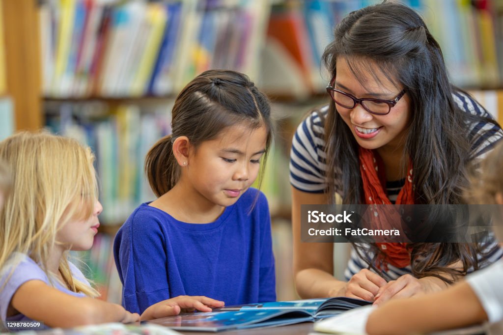 Thank you for helping us, teacher! A female teacher of Asian ethnicity is helping her multi ethnic group of students with a book to read. They are all dressed casually and are at their school library. Literacy Stock Photo