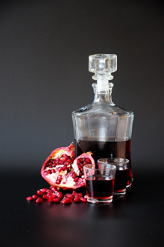 A decanter and two glasses of pomegranate liqueur next to the fruit and fruit seeds on a black background. Close-up.