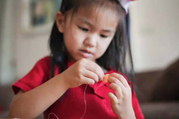 Photo of Close up hand of asian child girl is threading beads onto a string with intention and fun in home.