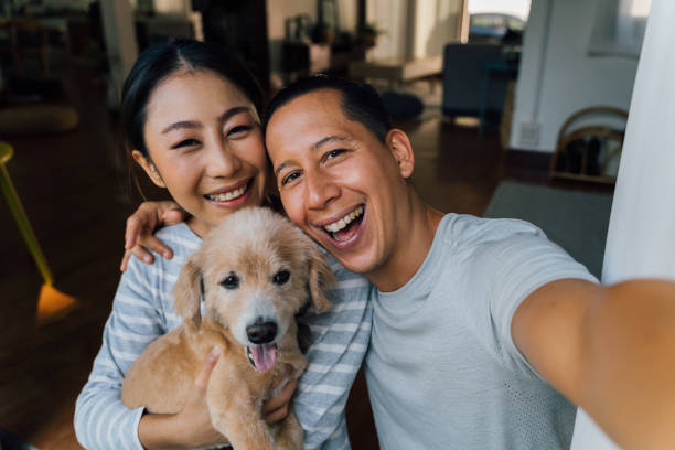 jovem casal asiático adulto segurando um cachorrinho tirando uma selfie de um telefone com interior doméstico no fundo. homem e mulher maduros dos anos 30 com animal de estimação tirando fotos de família. - retrato de grupo feliz. - pets family dog asian ethnicity - fotografias e filmes do acervo