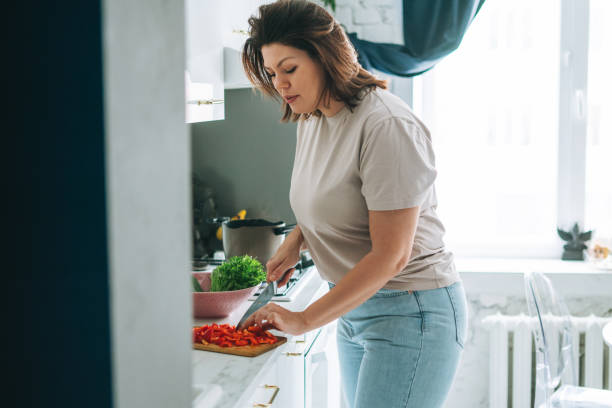 Beautiful brunette young woman plus size body positive cooking salad in kitchen at the home Beautiful brunette young woman plus size body positive cooking salad in kitchen at home woman making healthy dinner stock pictures, royalty-free photos & images