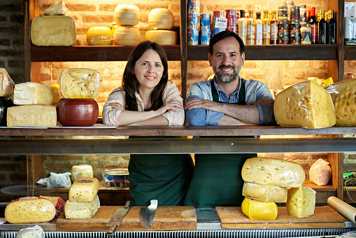 Partial front view of male and female business partners wearing aprons and standing side by side with arms crossed leaning on cheese counter in gourmet store.