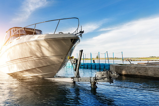 Big luxury cabin motorboat cruiser yacht launching at trailer ramp on river or lake. Warm morning sunrise sunshine reflection in calm water surface. Luxury rich fishing leisure recreation lifestyle.