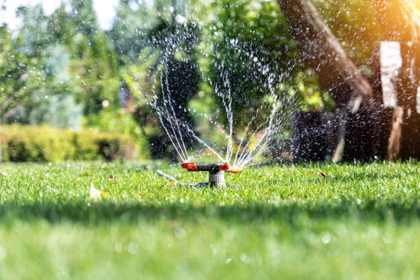 Landscape automatic garden watering system with different rotating sprinklers installed on turf. Landscape design with lawn and fruit garden irrigated with smart autonomous sprayers at sunset time stock photo