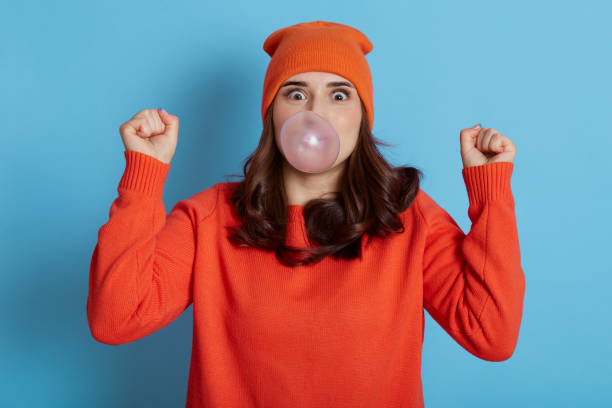istock Excited dark haired female blows bubble from chewing gum, has amazed expression, dresses casual clothes, model posing isolated over blue background, cant believe in her success, clenches fists. 1298838668
