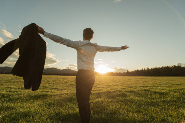 Businessman standing in beautiful green meadow Rear view of a businessman holding his suit standing in beautiful green meadow with his arms spread widely lit by the setting sun. prosperity stock pictures, royalty-free photos & images